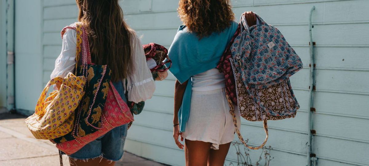 Two women walking away from the camera on a sidewalk, each carrying multiple patterned fabric backpacks and shoulder bags against a light blue wooden wall.
